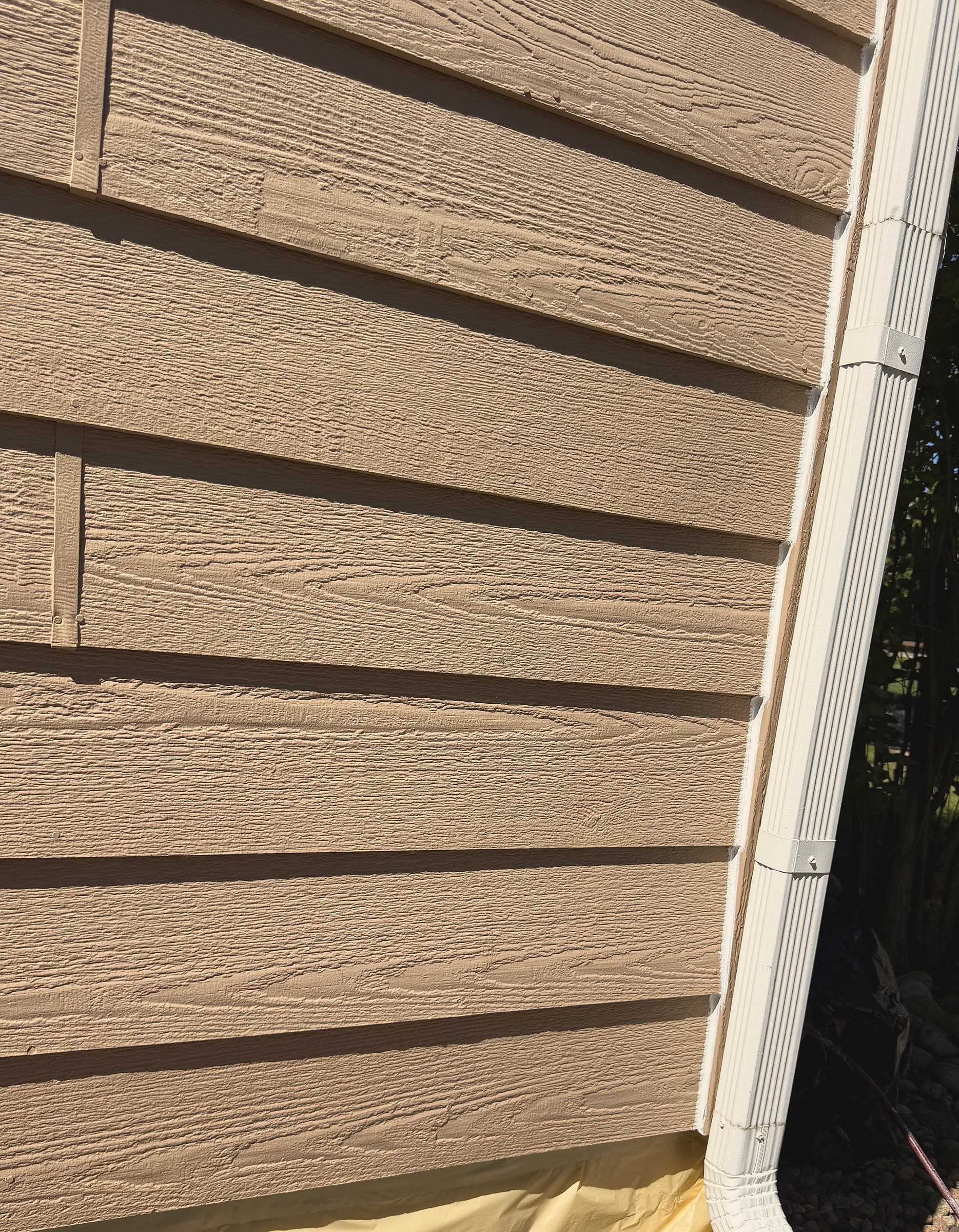 Tan lap siding and a white downspout after caulk and prep, with base masking that protects the house