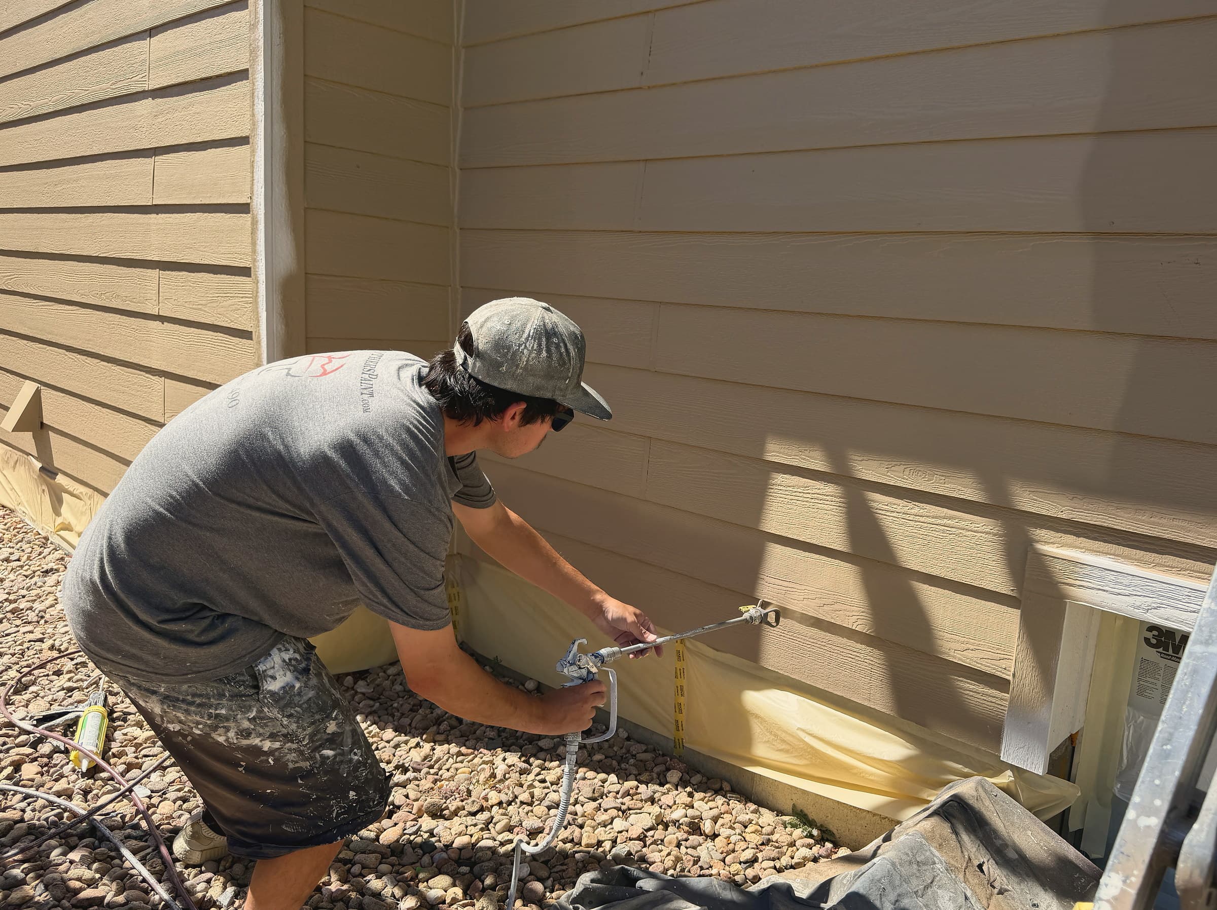 BrothersPaint.com painter spraying a tan lap-siding wall, with the foundation and a window masked